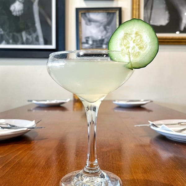 A light green cocktail in a coupe glass garnished with a cucumber slice on a wooden table.