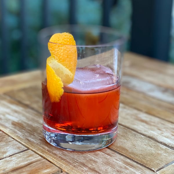 A vibrant red cocktail with a large ice cube and an orange peel garnish on a wooden table.