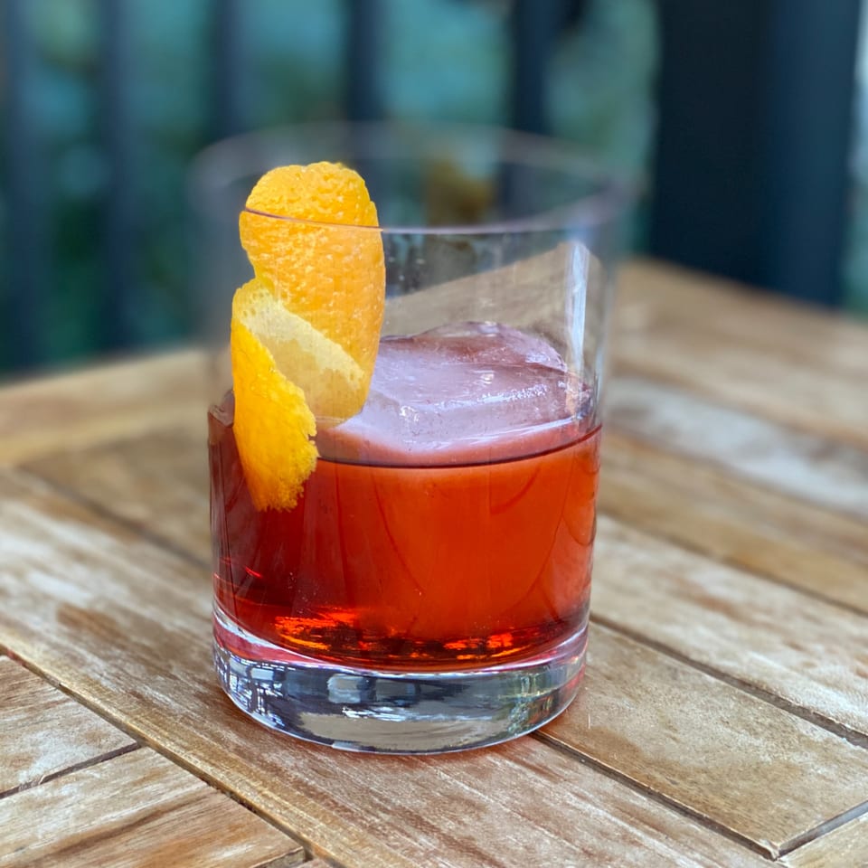 A vibrant red cocktail with a large ice cube and an orange peel garnish on a wooden table.