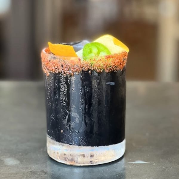 A dark cocktail with a chili-salted rim, garnished with orange and jalapeño slices.