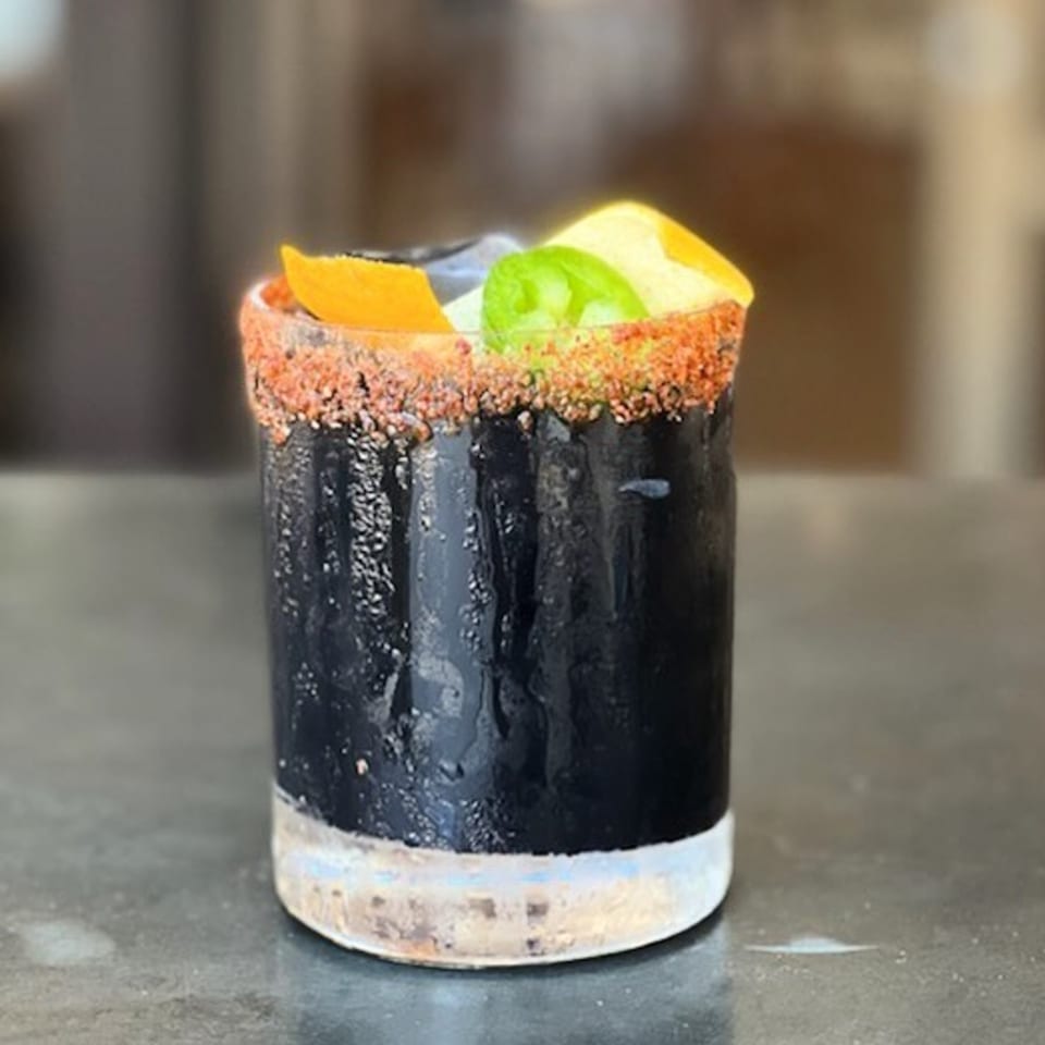 A dark cocktail with a chili-salted rim, garnished with orange and jalapeño slices.