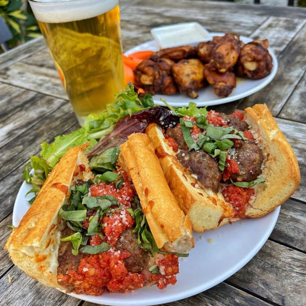 Meatball sub with salad, chicken wings, and a beer on a wooden table at The Shop: Pizza + Cocktails.