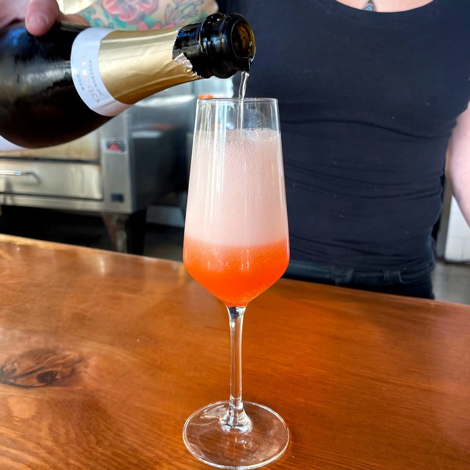 A person pours sparkling wine from a bottle into a glass with orange liquid on a wooden bar.
