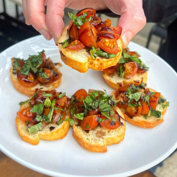 Hand holding a piece of bruschetta with tomatoes, basil, and balsamic glaze over a white plate.