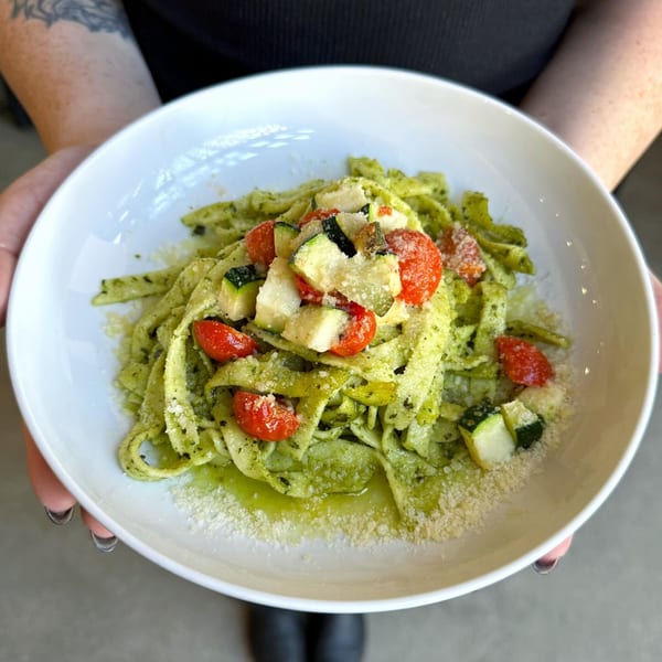 Hands holding a white bowl of green pesto pasta with cherry tomatoes, zucchini, and parmesan.