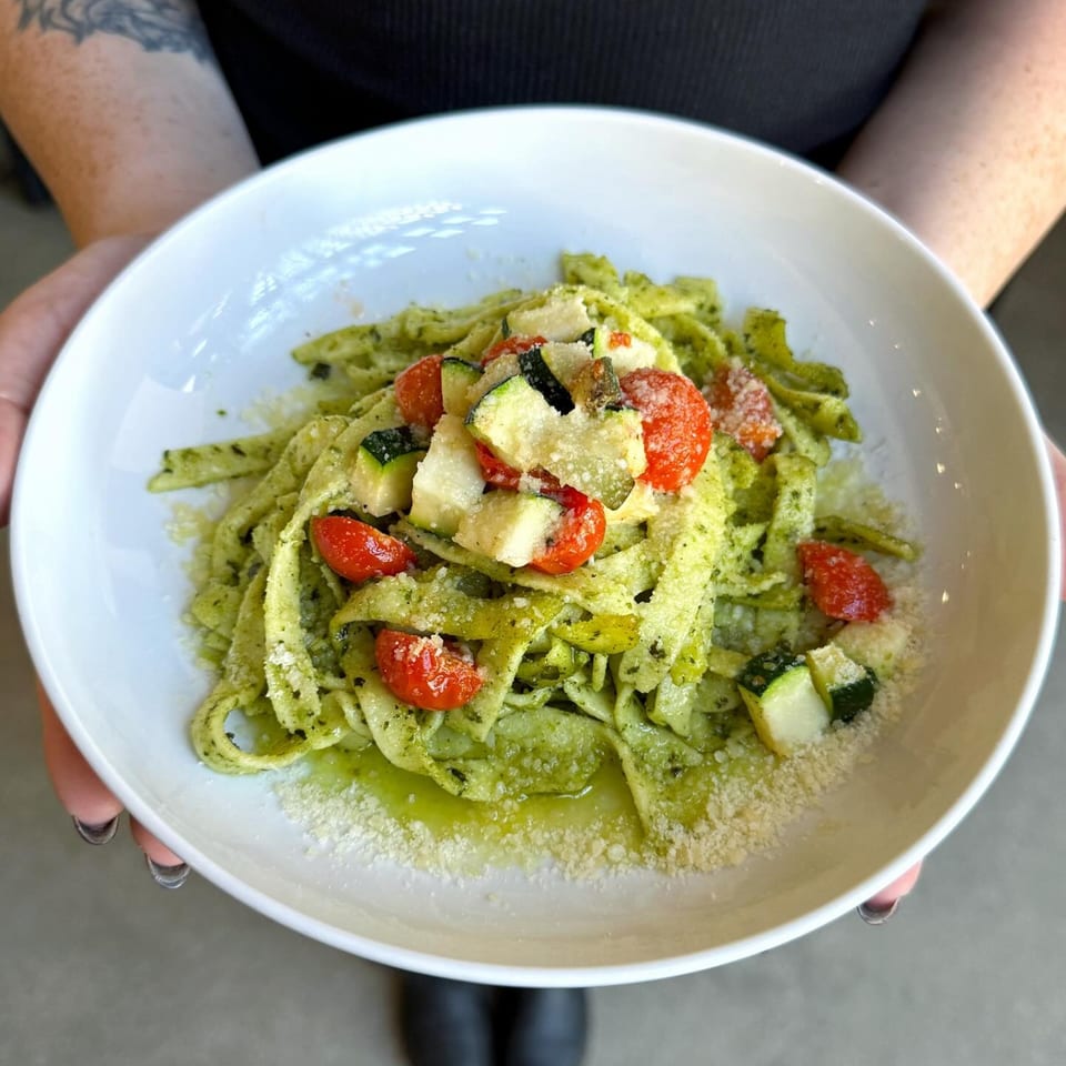 Hands holding a white bowl of green pesto pasta with cherry tomatoes, zucchini, and parmesan.