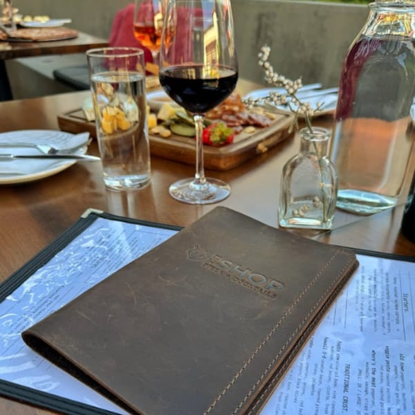 A leather menu for The Shop: Pizza + Cocktails rests on a table with drinks and food.