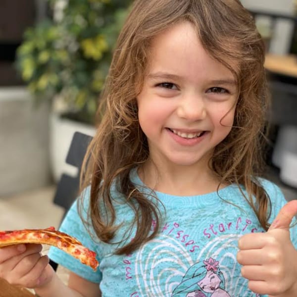 A smiling girl with long brown hair holds a slice of pizza and gives a thumbs up.