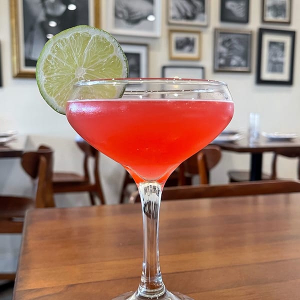 A vibrant red cocktail with a lime slice garnish sits on a wooden table at The Shop.