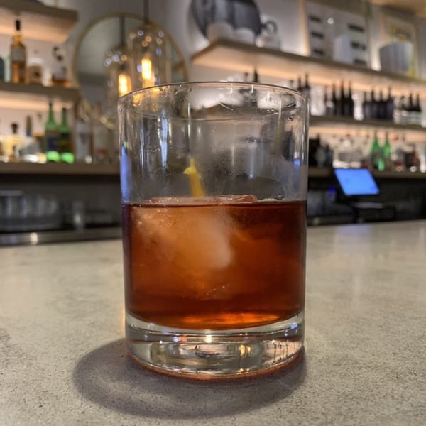 A cocktail with ice in a rocks glass on a bar counter, with liquor bottles blurred in the background.