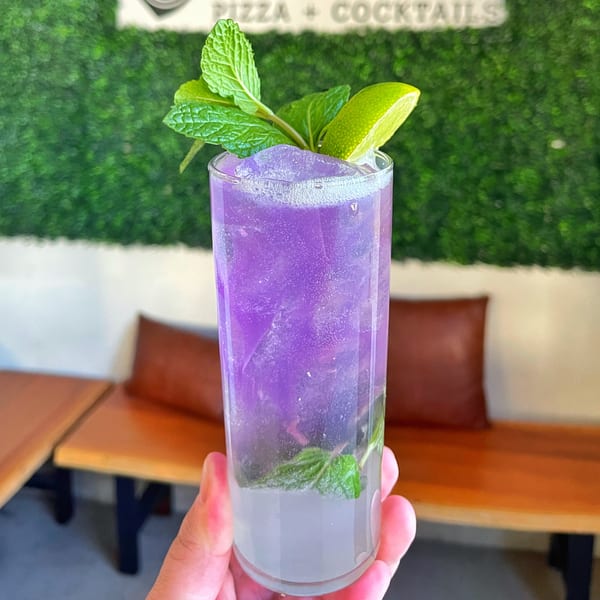 A hand holds a vibrant purple cocktail with mint and lime, against a green wall.