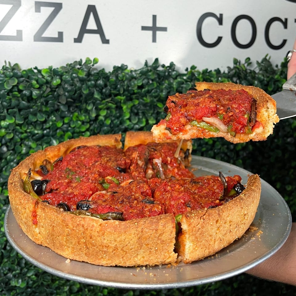 A deep-dish pizza from The Shop with a slice being lifted, revealing rich tomato sauce and toppings.