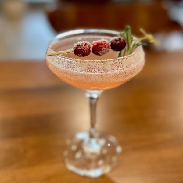 A festive cocktail with sugared cranberries and a rosemary sprig, served in a coupe glass.