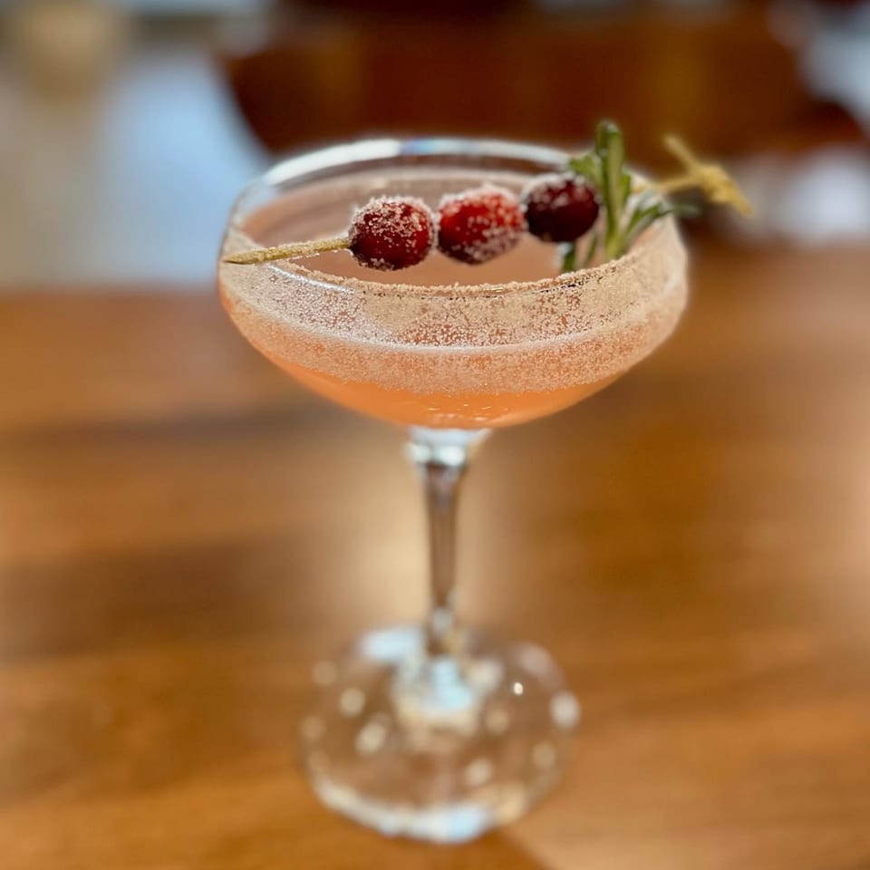 A festive cocktail with sugared cranberries and a rosemary sprig, served in a coupe glass.