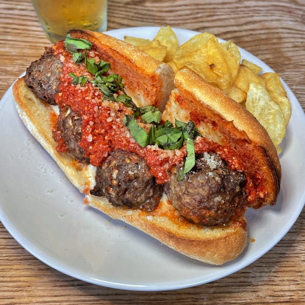 Meatball sub with red sauce, parmesan, and basil, served with potato chips and a drink.