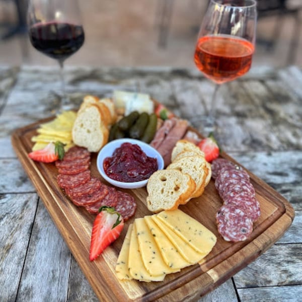 A wooden board with charcuterie, cheeses, bread, strawberries, and jam, with two wine glasses.